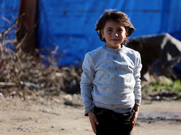 A young girl poses in front of a blue refugee tent.