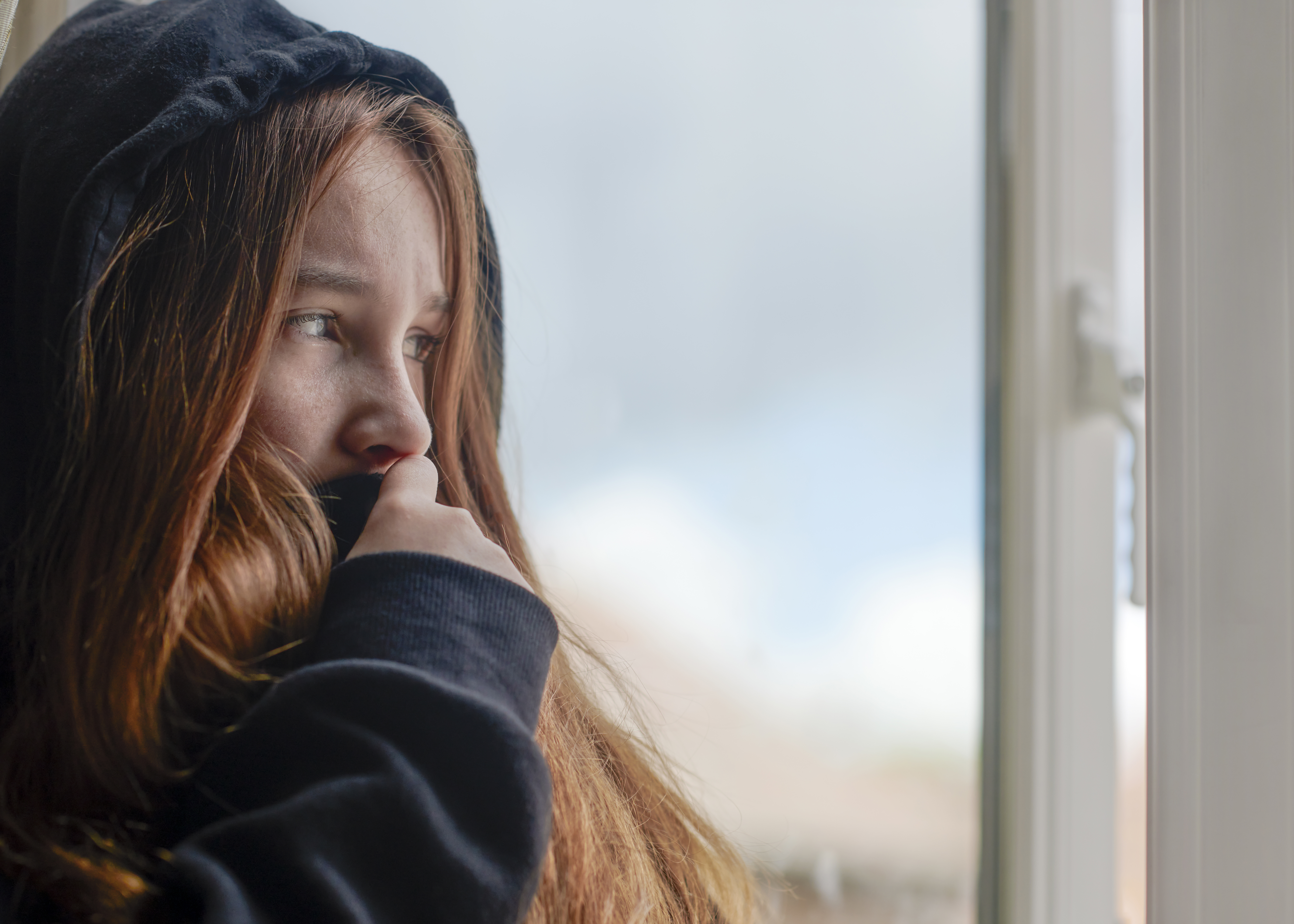 A teenage girl with red hair and wearing a gray hoodie looks out a window. 