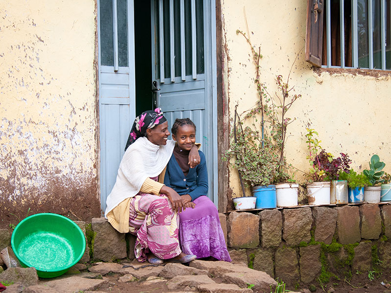 ethiopian woman hugs her teenage daughter as they sit on the steps outside their home