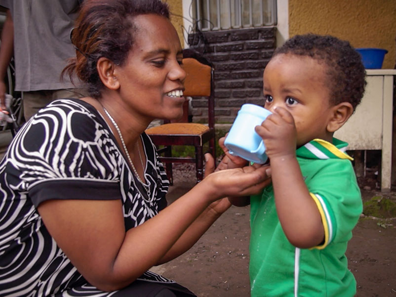 ethiopian mother helps her young son drink from a mug outside their home