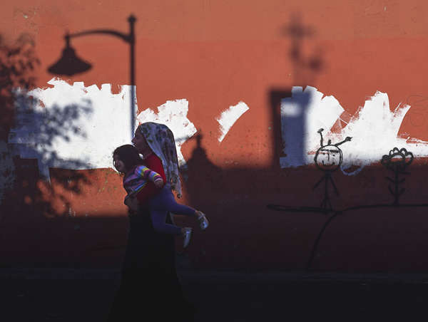 A profile view of a  refugee mom carrying her young children past a graffitied wall.
