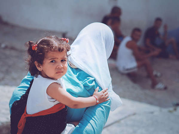 A mother with her toddler daughter on her back walks toward a group of refugees outside a building as the child looks back toward the direction they came from.