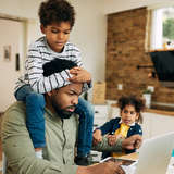 stressed Black foster dad searches for help on his laptop while kids tug on his arm