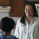 Asian foster mother talks to young refugee boy in kitchen
